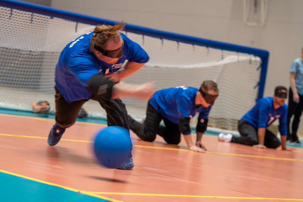Sinisessä pelipaidassa pelaava maalipalloilijamies juuri pallon heittäneenä. Taustalla joukkueen muita pelaajia maalin edustalla.